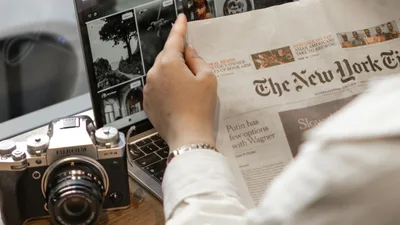 Close-up of a person reading The New York Times by a laptop and camera.